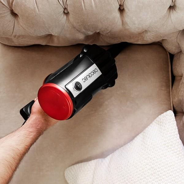Una mano che tiene un aspirapolvere portatile nero e rosso sopra un cuscino del divano beige.