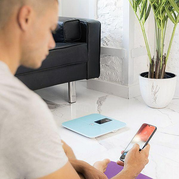 Hombre con teléfono cerca de una báscula azul. Sofá negro, suelo blanco y planta verde de fondo.