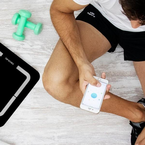 Osoba trzyma telefon, patrząc na ekran. W scenie waga i hantle.