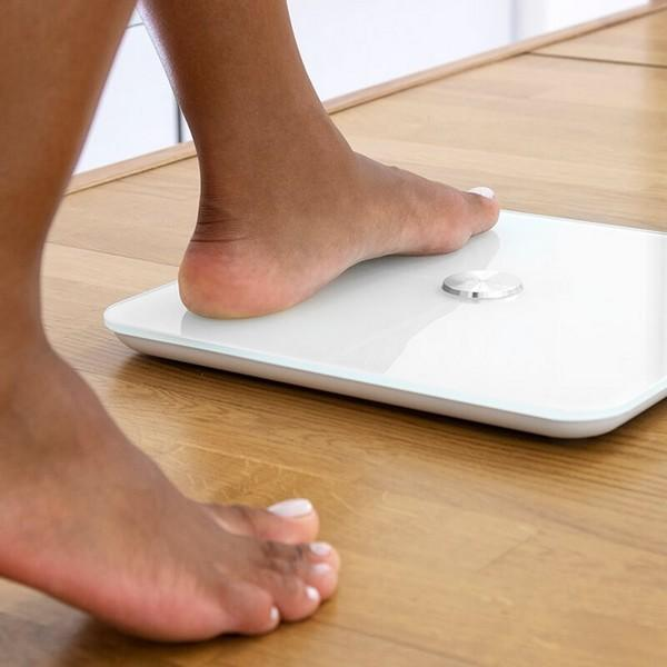 Die Füße einer Person auf einer weißen Digitalwaage auf einem Holzboden. Ein Fuß ist drauf, der andere nicht.