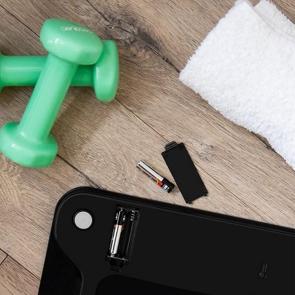 Czarna waga z bateriami, zielone hantle i biały ręcznik na drewnianej podłodze.