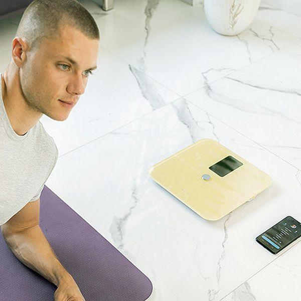 Mężczyzna stojący obok wagi, patrzący na telefon. Marmurowa podłoga, mata do jogi.