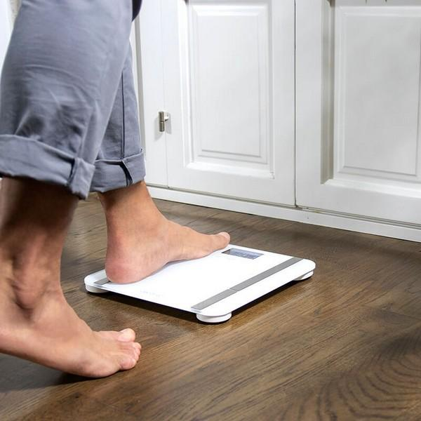 Osoba wchodzi na białą wagę na drewnianej podłodze, w pobliżu białych drzwi.