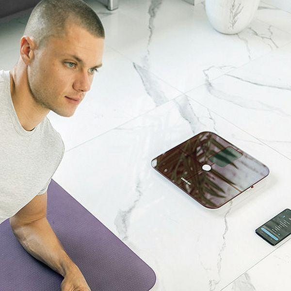 Ein Mann steht neben einer Waage. Eine Matte und ein Telefon sind ebenfalls sichtbar. Der Hintergrund hat Marmor.