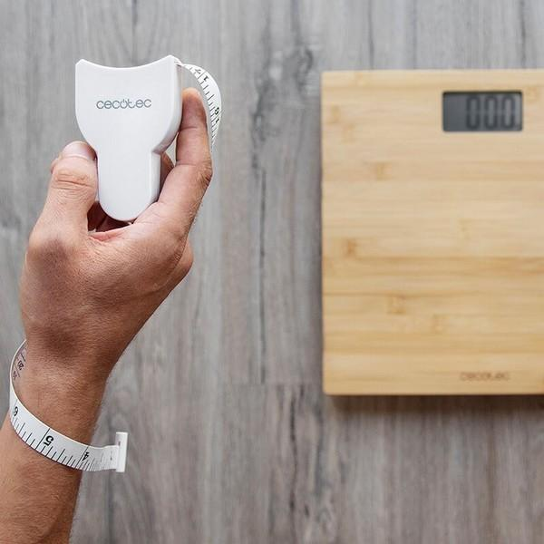 Osoba trzymająca białą miarkę. W tle znajduje się drewniana waga. Tłem jest drewniana podłoga.