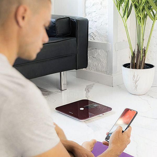 Mann mit Telefon neben Waage, mit Pflanze und Couch im Hintergrund.