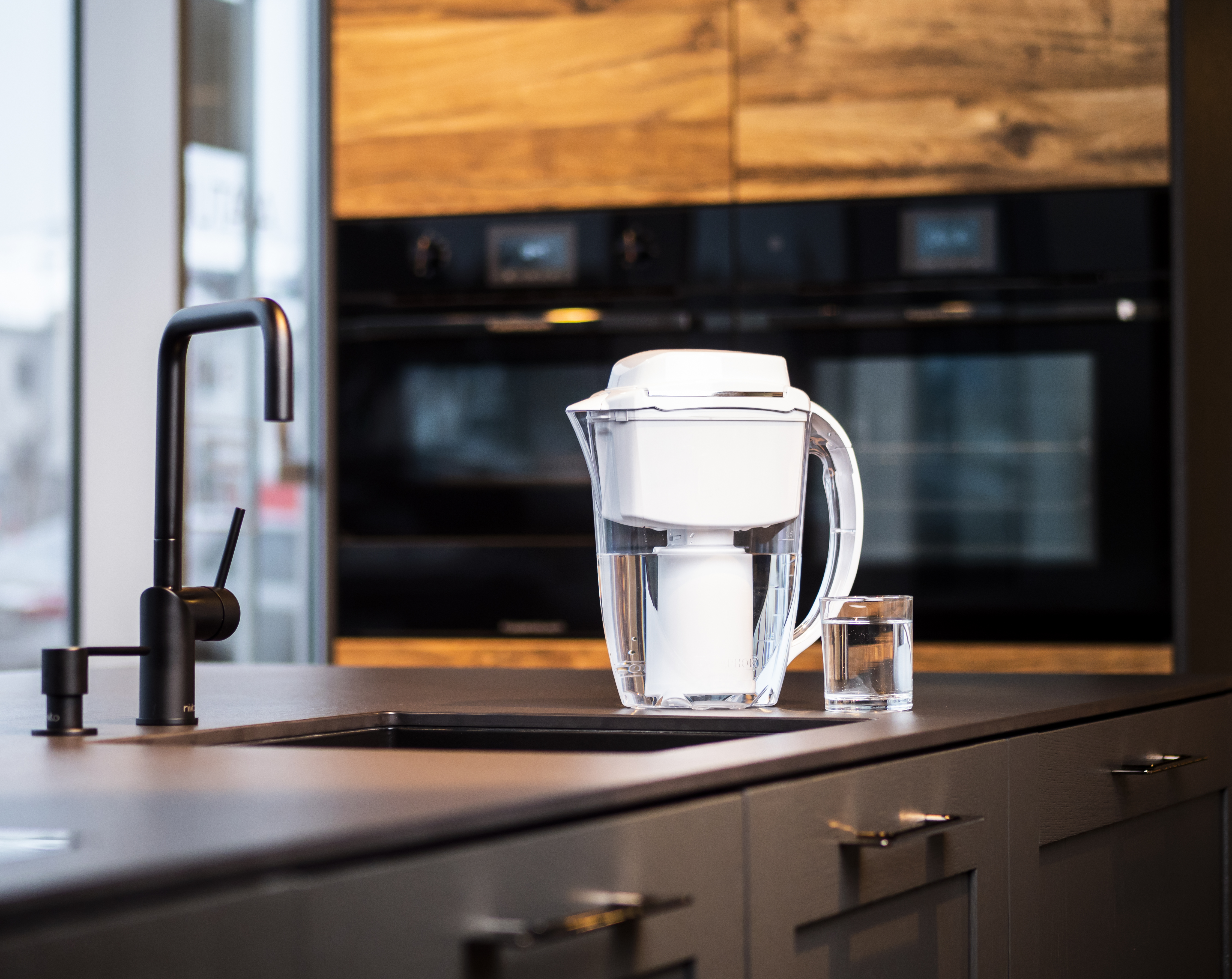 Ein weißer Wasserfilter-Krug auf einer dunklen Arbeitsplatte mit Wasserhahn und einem Glas Wasser.