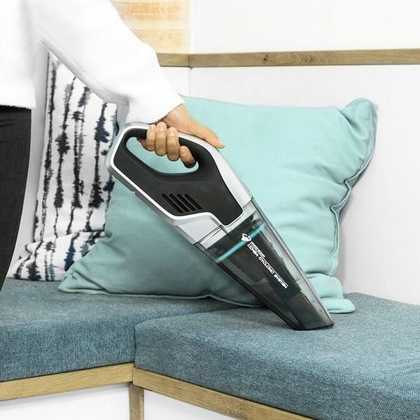 Eine Hand, die einen Handstaubsauger über einem blauen Kissen hält, mit einem türkisfarbenen Sofa im Hintergrund.