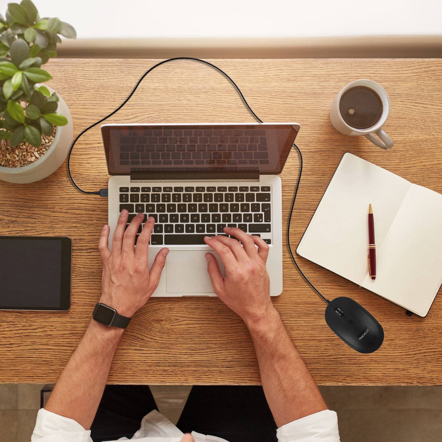 Eine Person, die auf einem Laptop mit Maus, Notizblock und Kaffeetasse tippt. Der Schreibtisch ist aus Holz.
