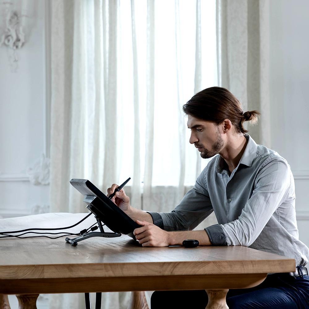 Mann, der mit einem Stift auf einem Tablet an einem Holztisch zeichnet. Der Hintergrund ist ein Raum.