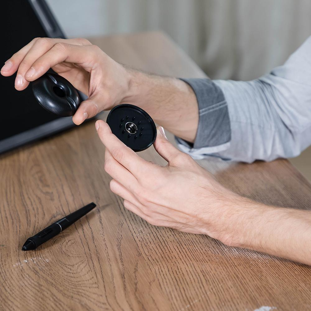 Hände, die schwarze Tech-Teile auf einem Holztisch halten und zusammensetzen. Vordergrund: Teile eines Geräts.