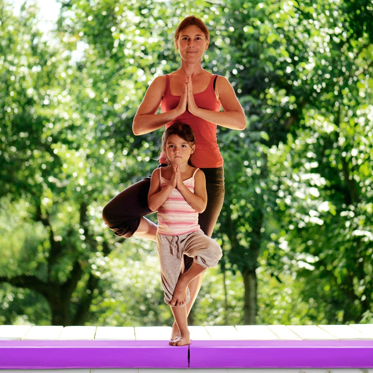 Frau und Mädchen machen eine Yoga-Pose. Sie stehen auf einer lila Matte im Freien mit Bäumen im Hintergrund.
