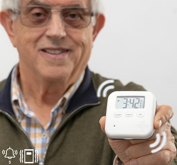 Ein älterer Mann hält einen weißen Digitaltimer und lächelt in die Kamera. Weißer Hintergrund.