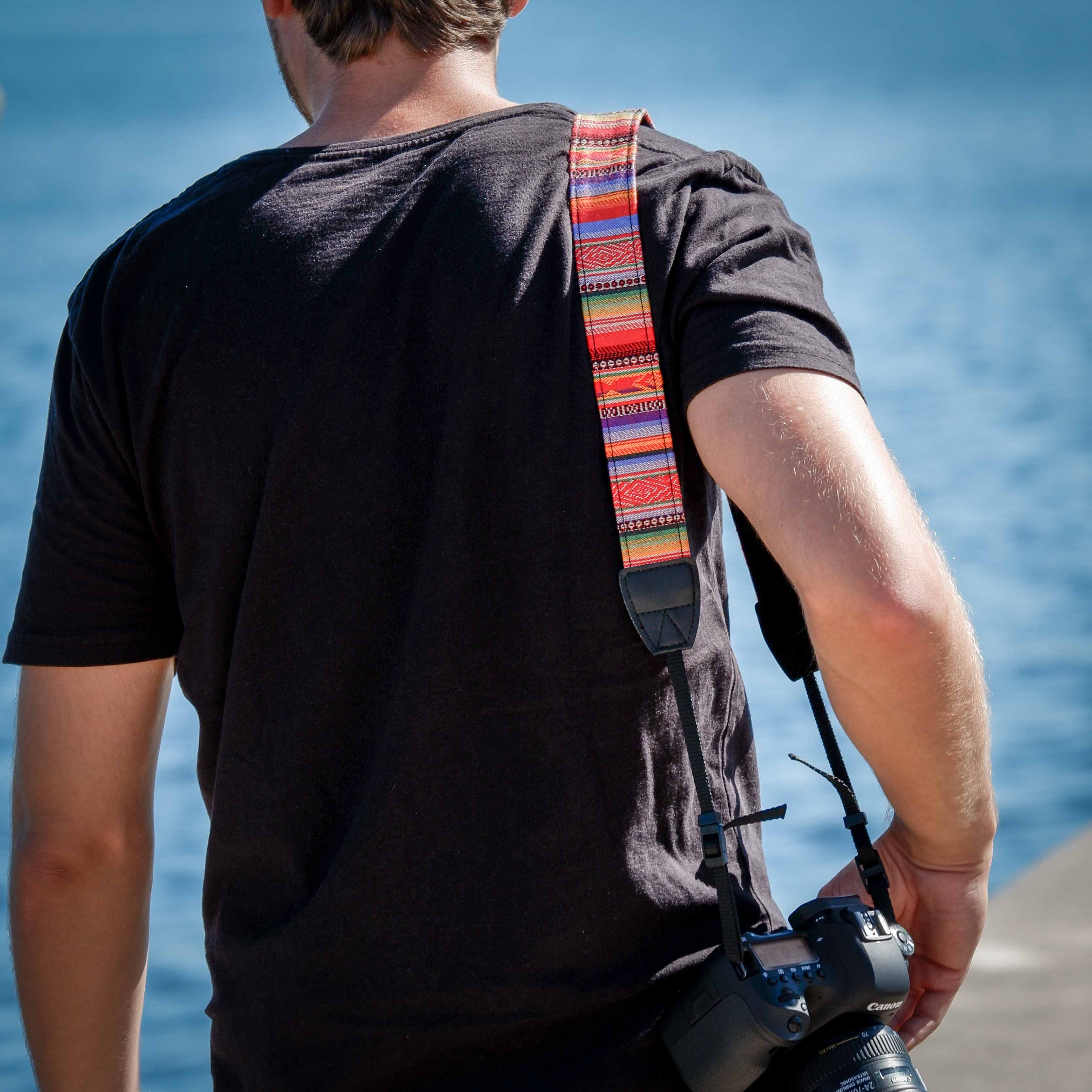 Mann mit schwarzem Hemd und Kamera, bunter Gurt. Im Hintergrund ist das Meer zu sehen.