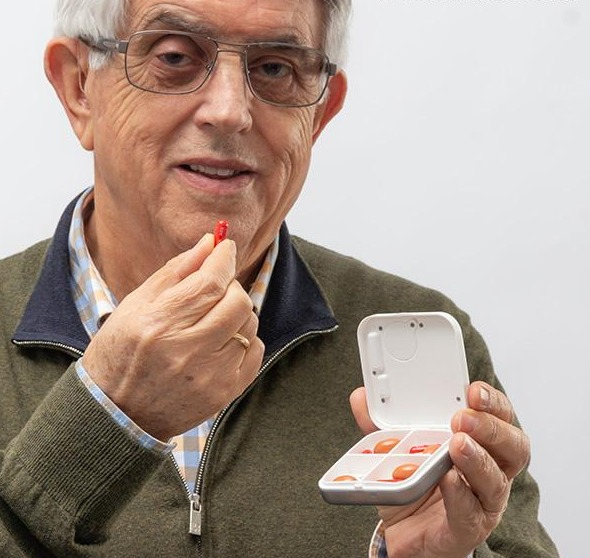 Ein älterer Mann hält eine Pille und einen Pillenorganizer. Er trägt eine Brille und einen grünen Pullover.