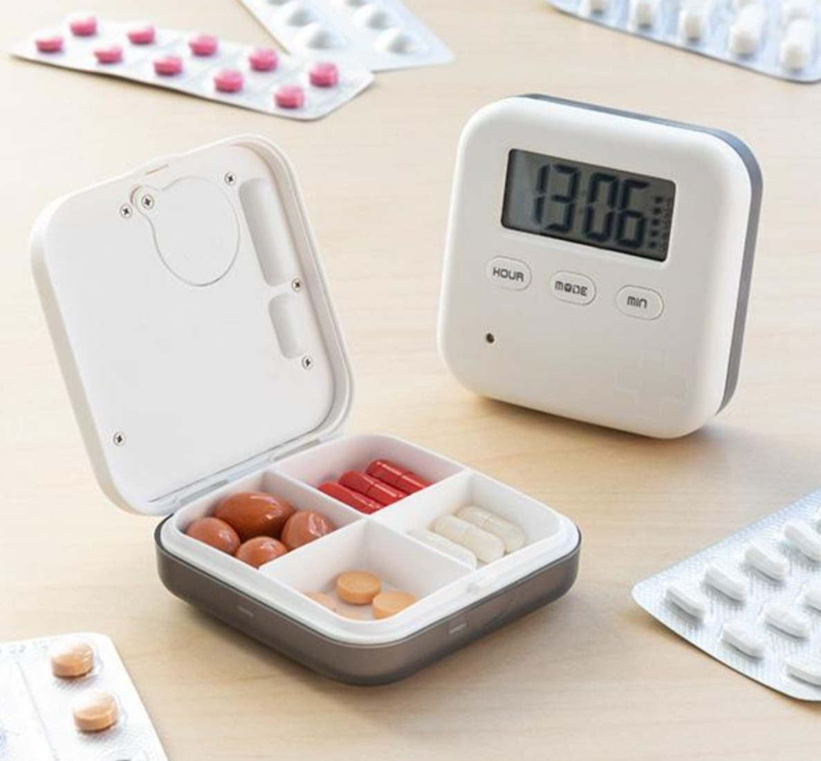 Ein Tabletten-Organizer mit digitaler Uhranzeige. Er ist geöffnet und zeigt Tabletten. Daneben Blisterpackungen.