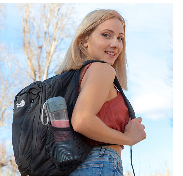 Kobieta z plecakiem uśmiecha się do kamery, trzymając butelkę smoothie.