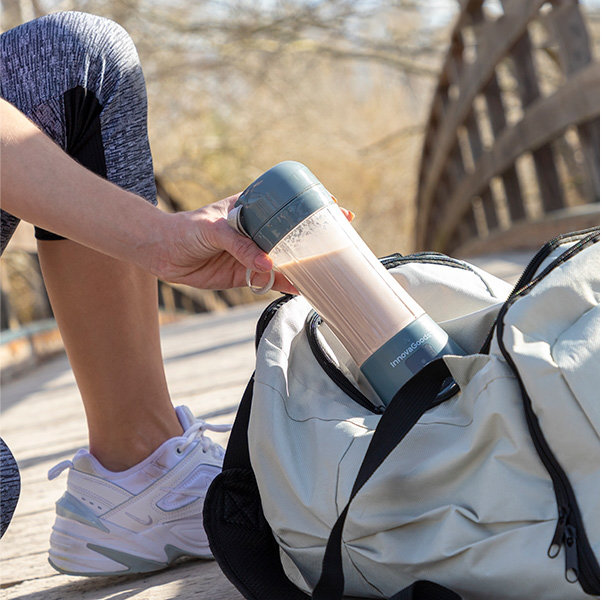 Osoba wkładająca butelkę blendera z płynem do szarej torby sportowej, na zewnątrz.