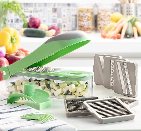 Tritatutto per verdure verde con lame e un contenitore pieno di verdure tritate, vari frutti sullo sfondo.