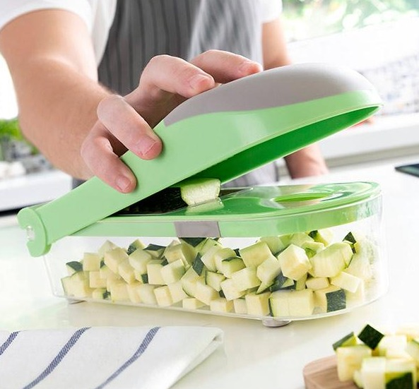 Una persona taglia zucchine con un tritatutto verde e trasparente in un contenitore trasparente. Le zucchine sono a cubetti.