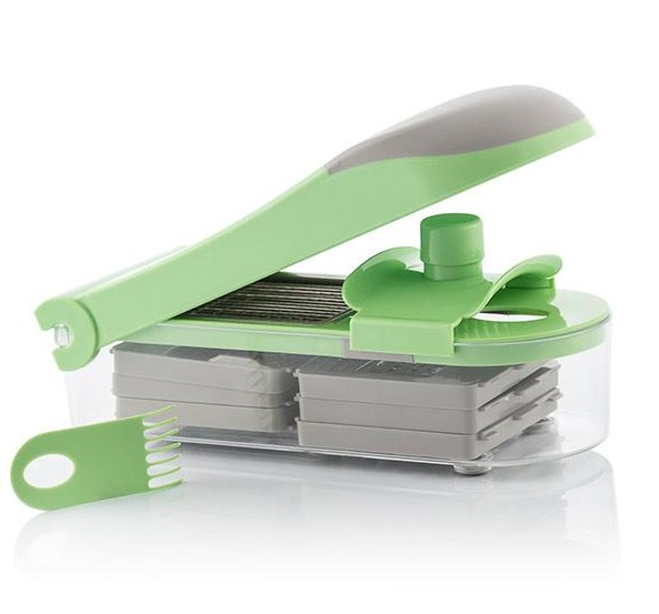 Affettatrice per verdure verde e grigia con diverse lame e un contenitore trasparente. È su una superficie bianca.