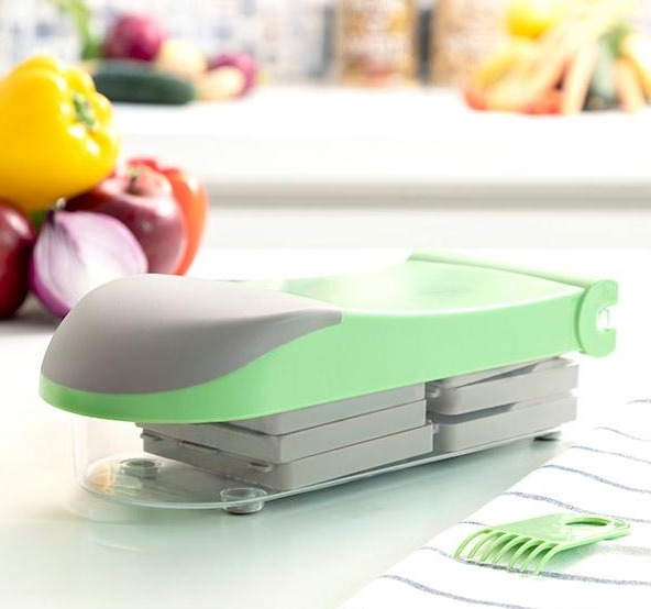Un tritatutto per verdure verde e grigio su una superficie bianca. Ci sono verdure sullo sfondo.
