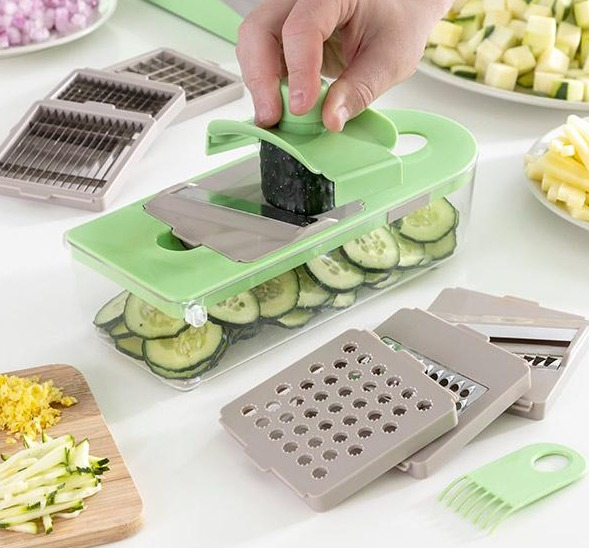Affettatrice di verdure verde con una mano che la usa. Cetrioli a fette e altri accessori da taglio sono visibili.