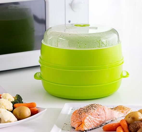 Vaporera de microondas verde con salmón y verduras, platos de comida y microondas al fondo.