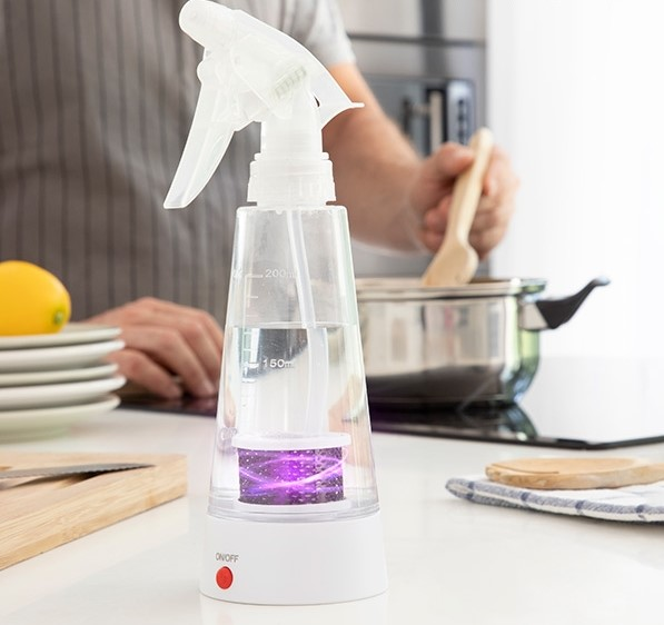 Una persona rociando líquido de una botella de spray transparente. En el fondo, una persona revuelve comida en una olla.
