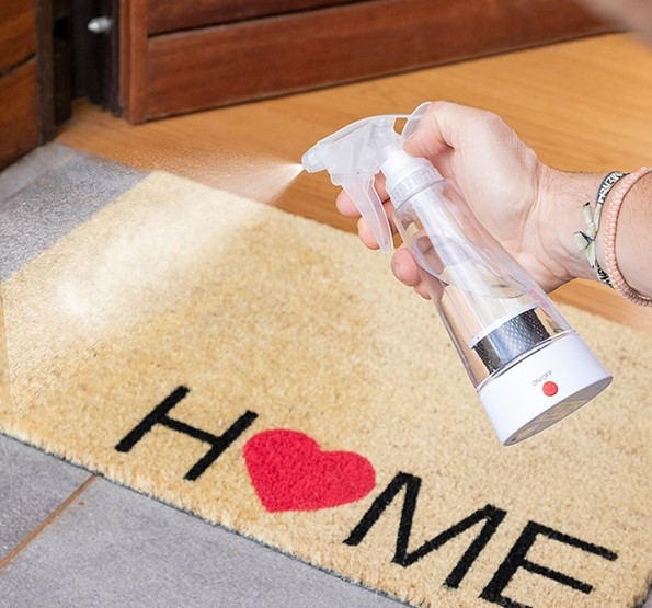 Mano rociando una alfombra de casa con un pulverizador. La alfombra tiene un corazón rojo y la palabra HOME.