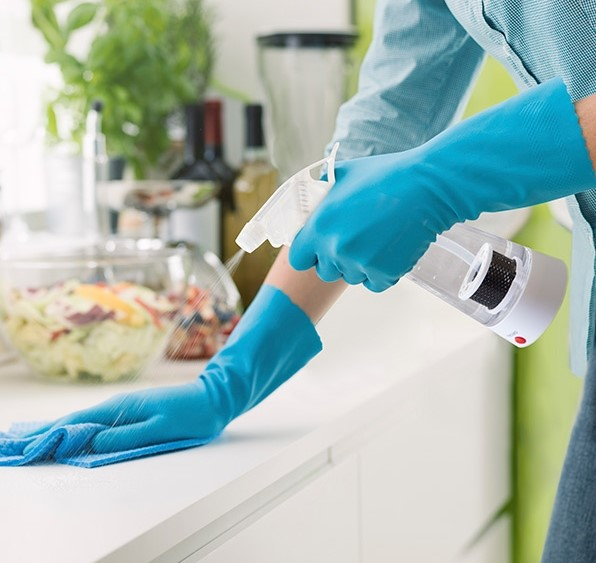Persona con guantes azules rociando limpiador sobre una superficie blanca en la cocina.