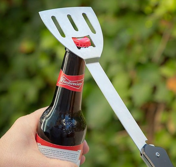 Ein Pfannenwender mit Flaschenöffner öffnet eine Bierflasche. Die Flasche hat ein Budweiser-Etikett.