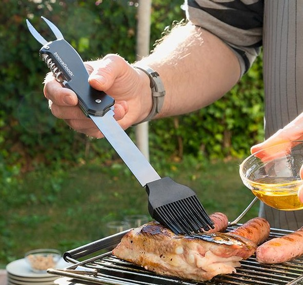 Eine Person grillt, benutzt ein Werkzeug mit Pinsel- und Gabelmerkmalen. Fleisch und Würstchen auf dem Grill.