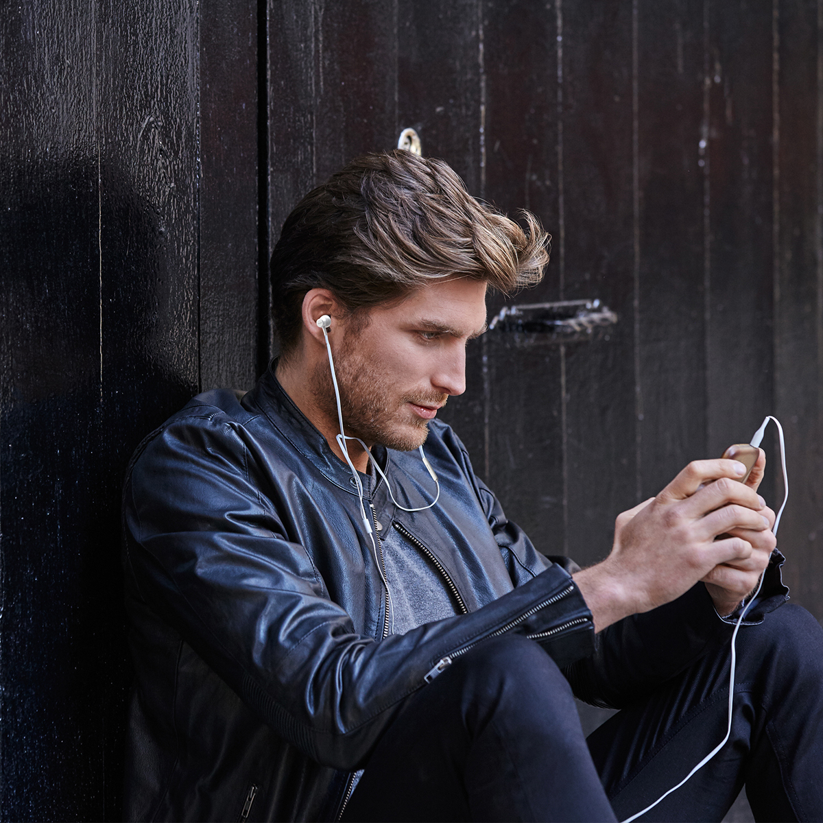 Mężczyzna siedzący, w skórzanej kurtce, słuchający muzyki z białymi słuchawkami.