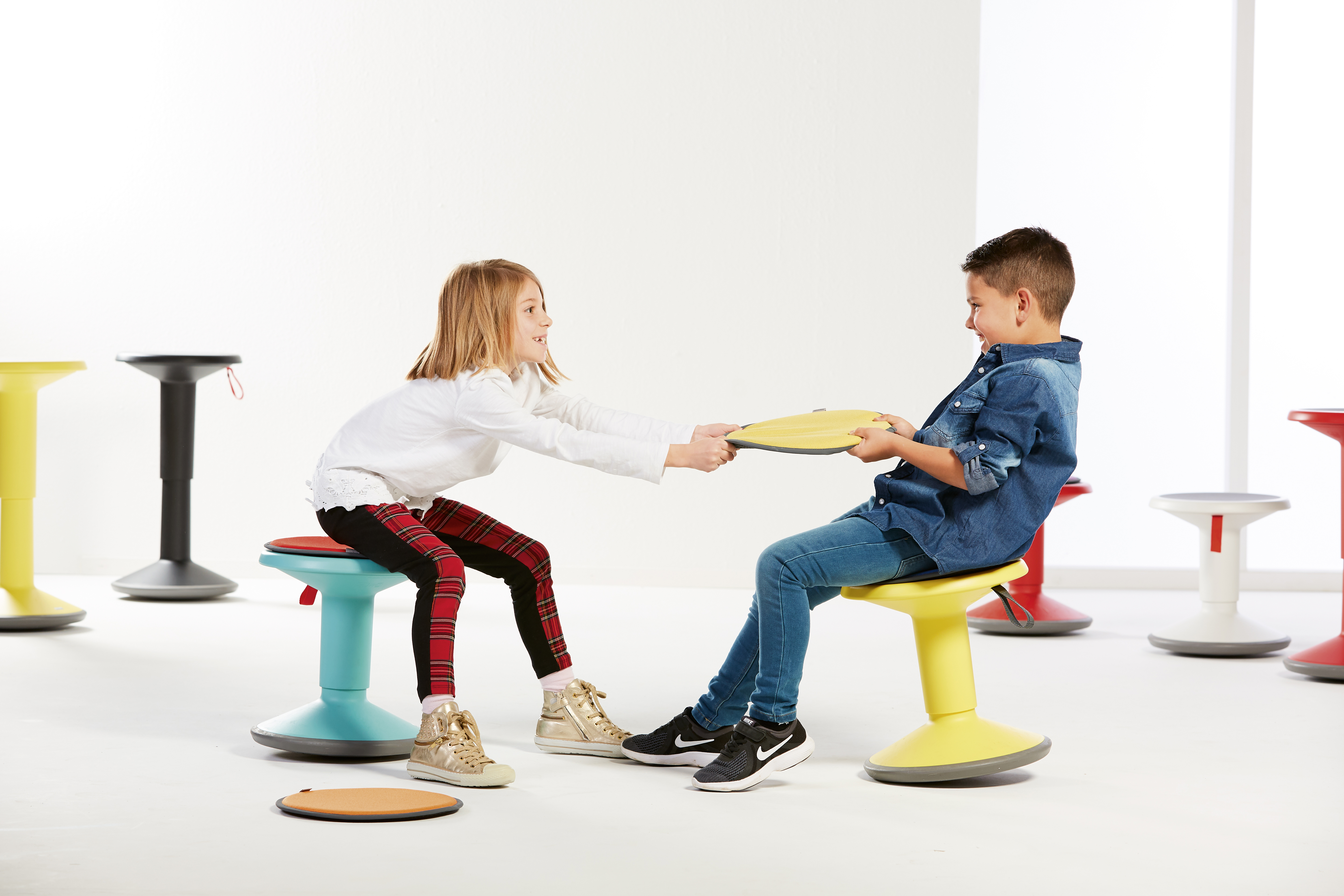 Zwei Kinder, ein Mädchen und ein Junge, ziehen an einer Scheibe, während sie auf bunten Hockern sitzen.