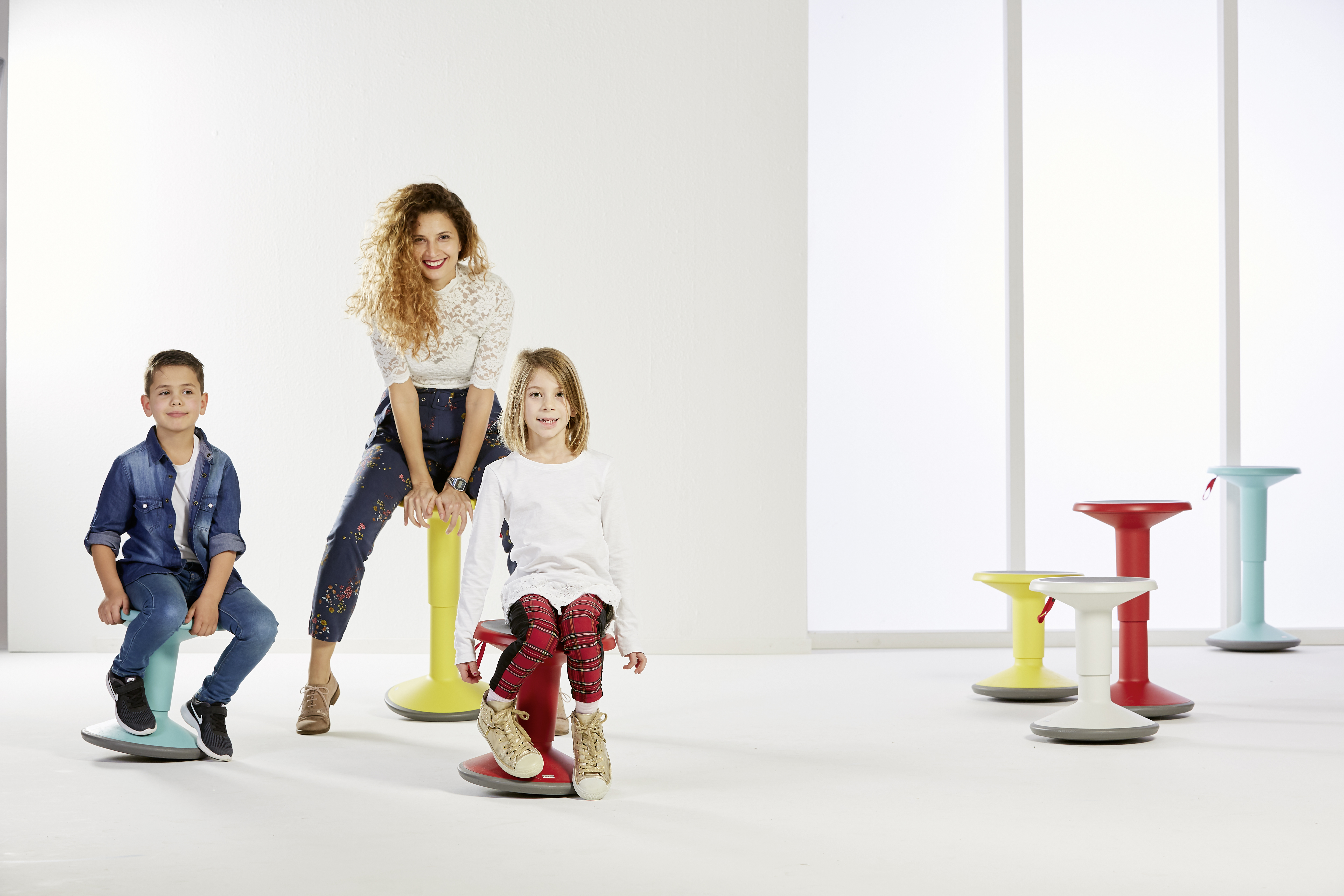Frau, zwei Kinder und mehrere Hocker. Die Kinder und die Frau sitzen auf Hockern. Weißer Hintergrund.