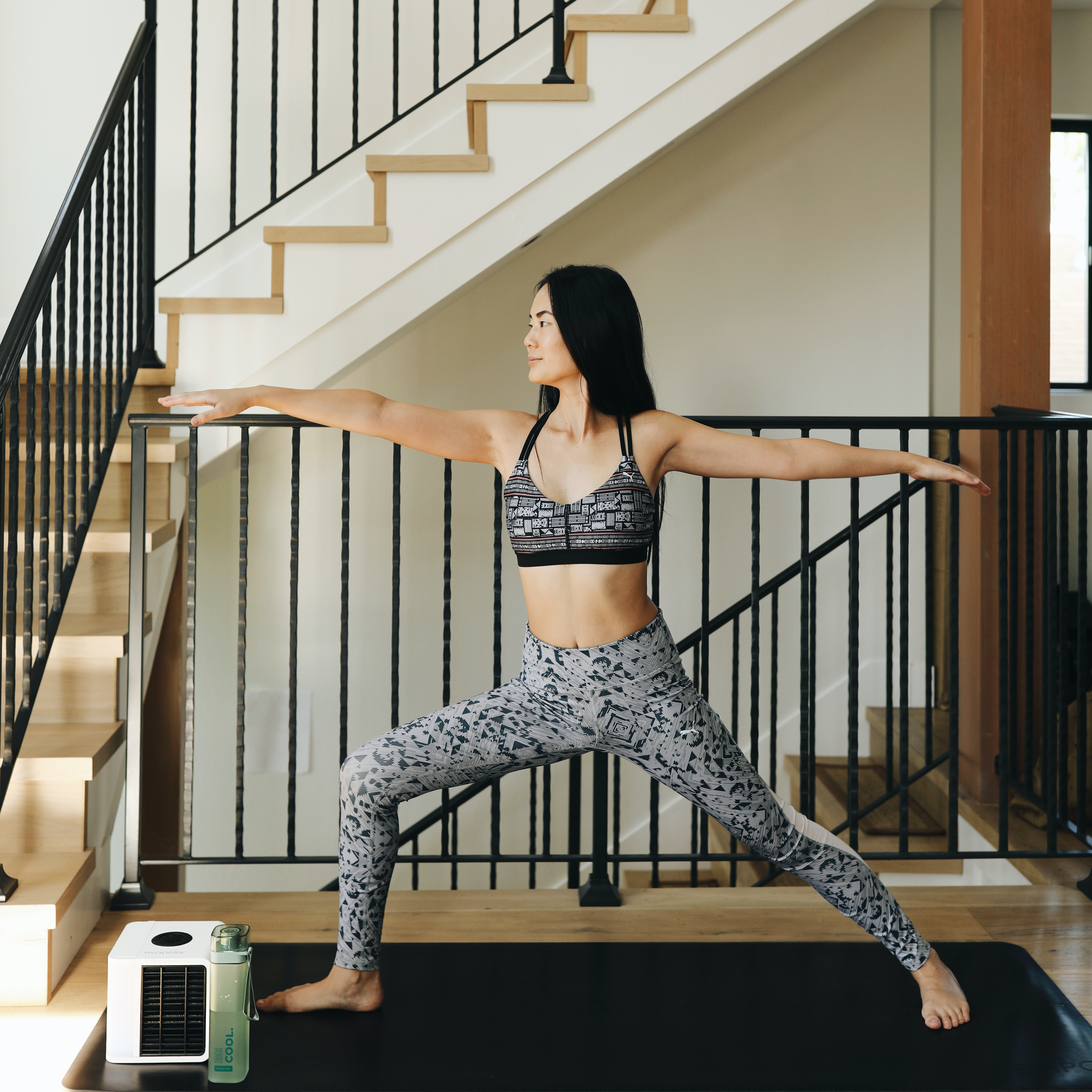 Frau in Sportkleidung macht eine Yoga-Pose. Schwarze Matte, Treppe und Luftkühler im Hintergrund.
