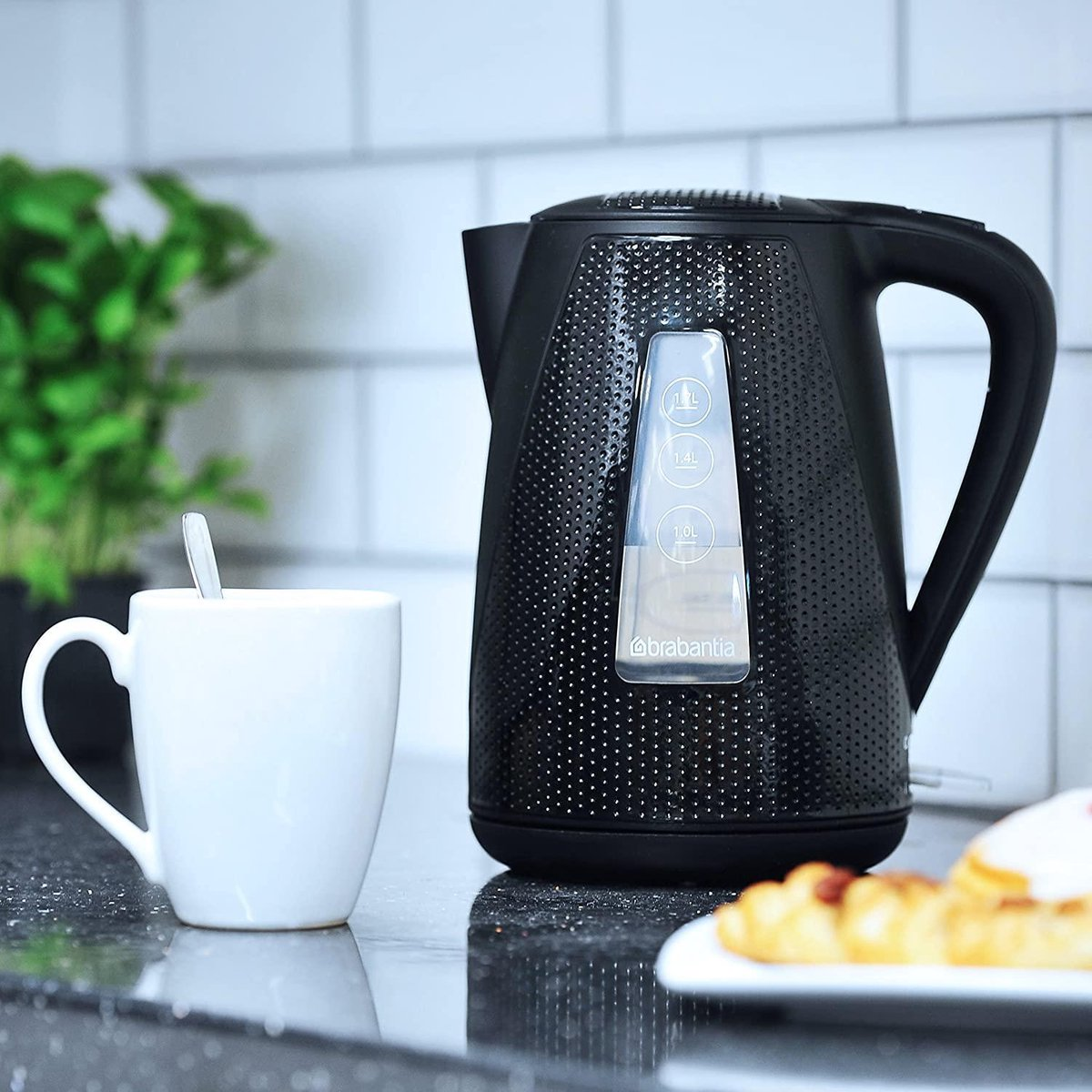 Zwarte elektrische waterkoker met doorzichtig waterniveauvenster. Witte mok en croissant op een wit bord. Keuken.