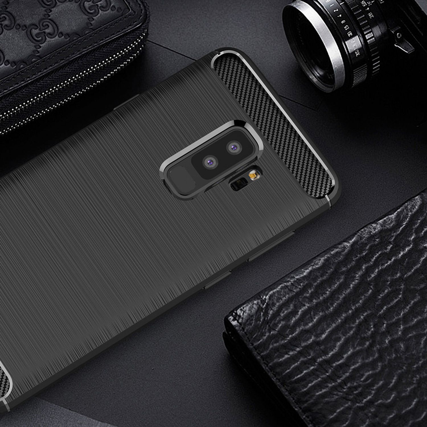 Eine schwarze Handyhülle mit Karbonfaser-Design, eine Geldbörse und eine Kamera auf dunkler Oberfläche.