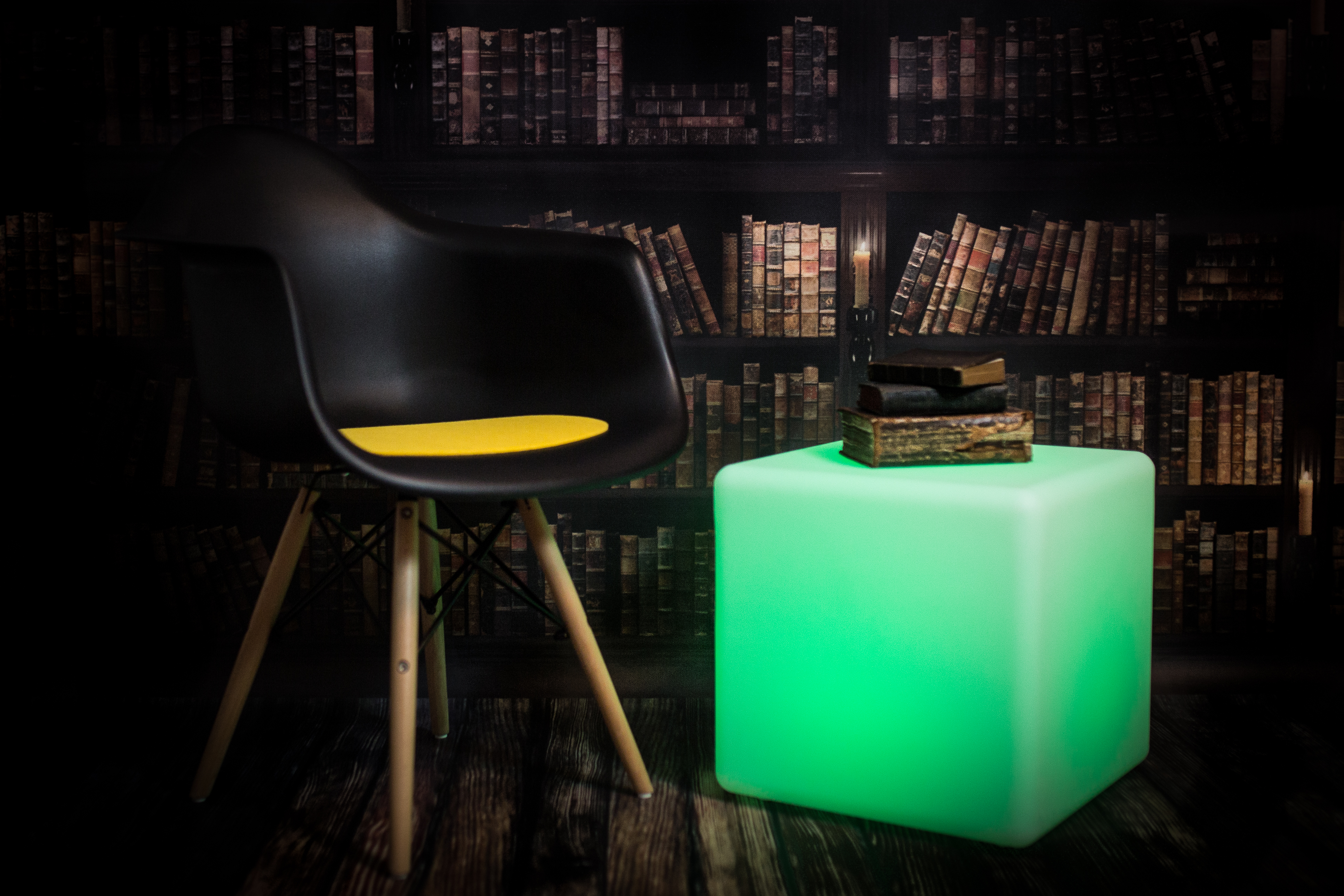 Ein schwarzer Stuhl, ein grün leuchtender Würfel und ein Stapel Bücher auf einem Tisch, im Hintergrund eine Bibliothek.