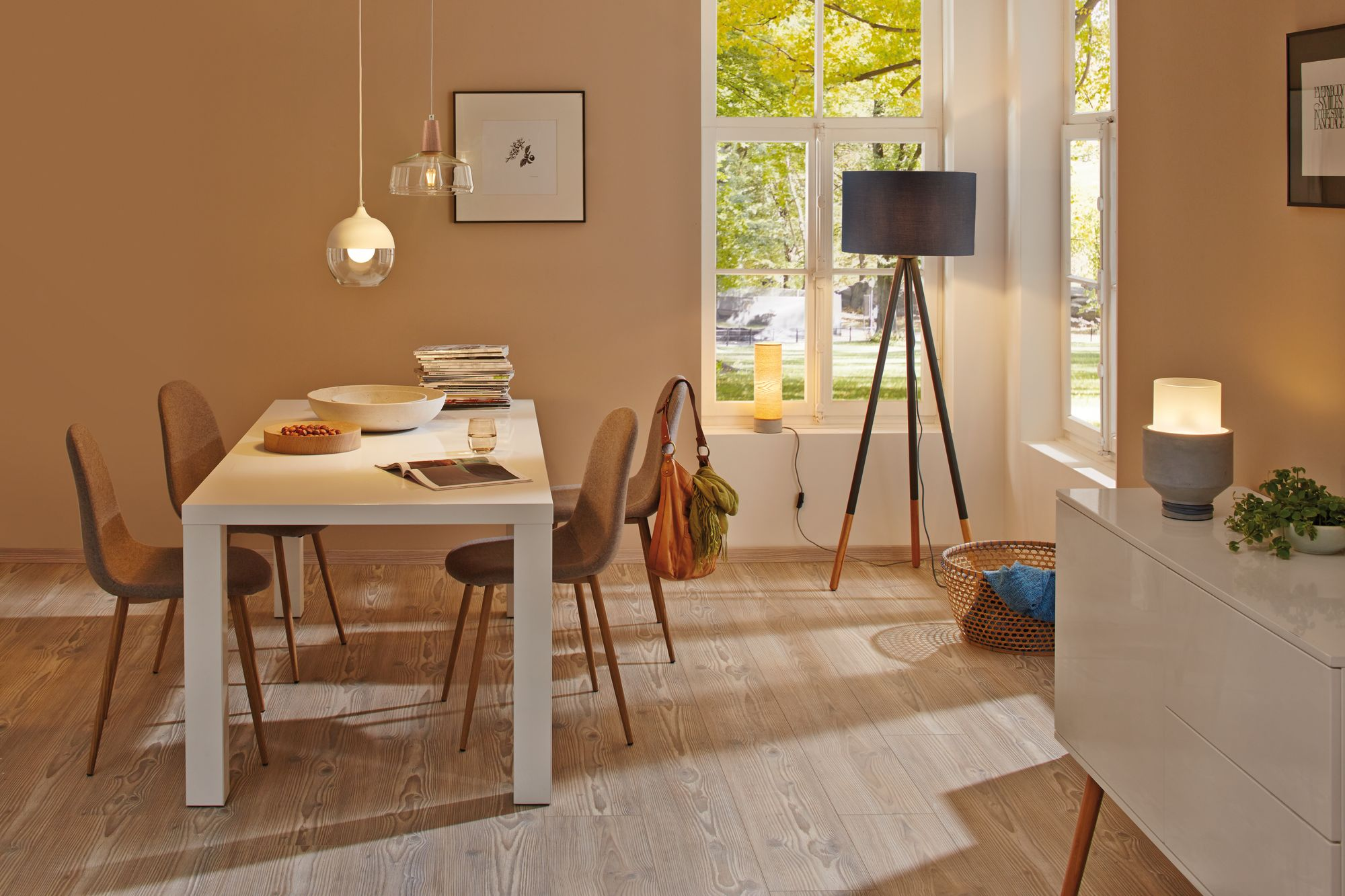 Esszimmer. Weißer Tisch mit Stühlen, zwei Hängelampen und eine Stehlampe. Helle Wände und Holzboden.