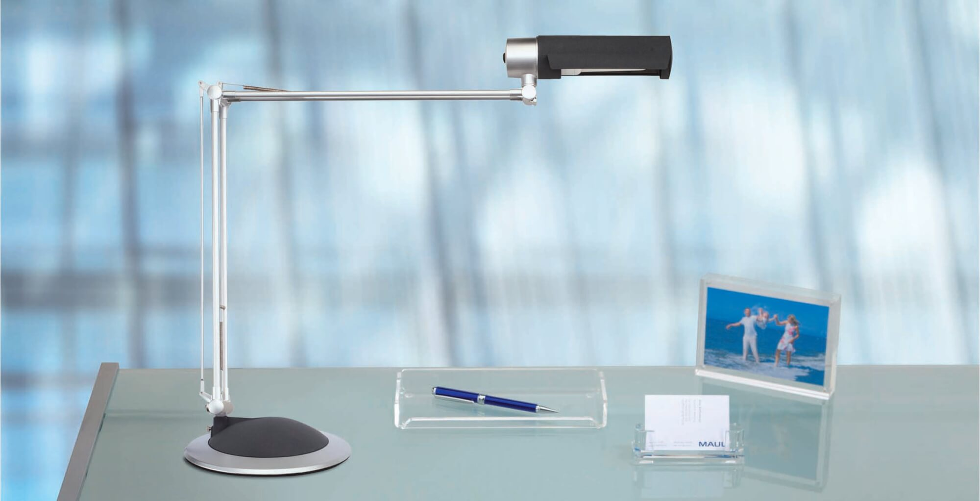 Lampa biurkowa, długopis, ramka na zdjęcia i wizytownik na stole.