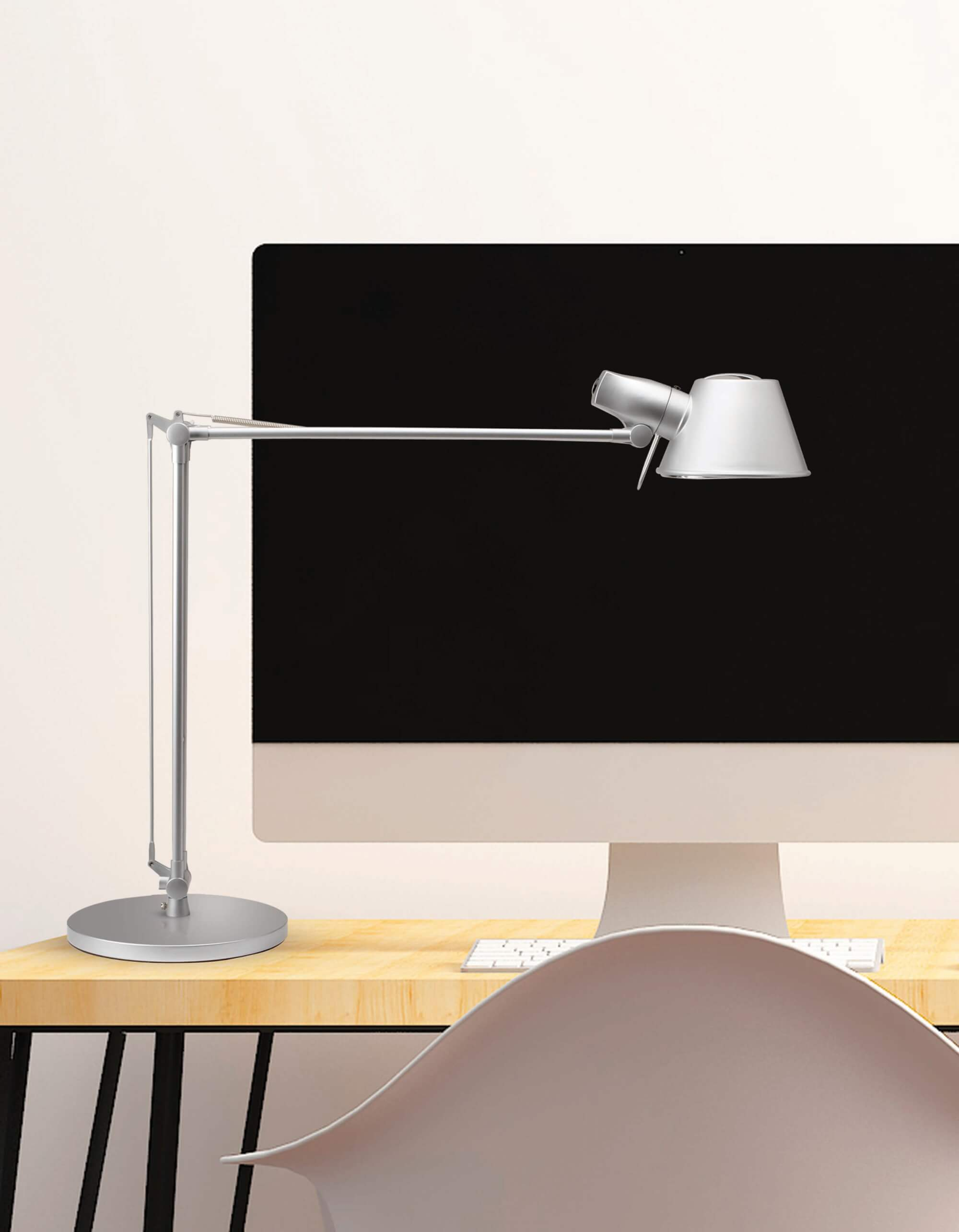 Srebrna lampa biurkowa skierowana w stronę monitora komputera na drewnianym biurku, biała ściana.