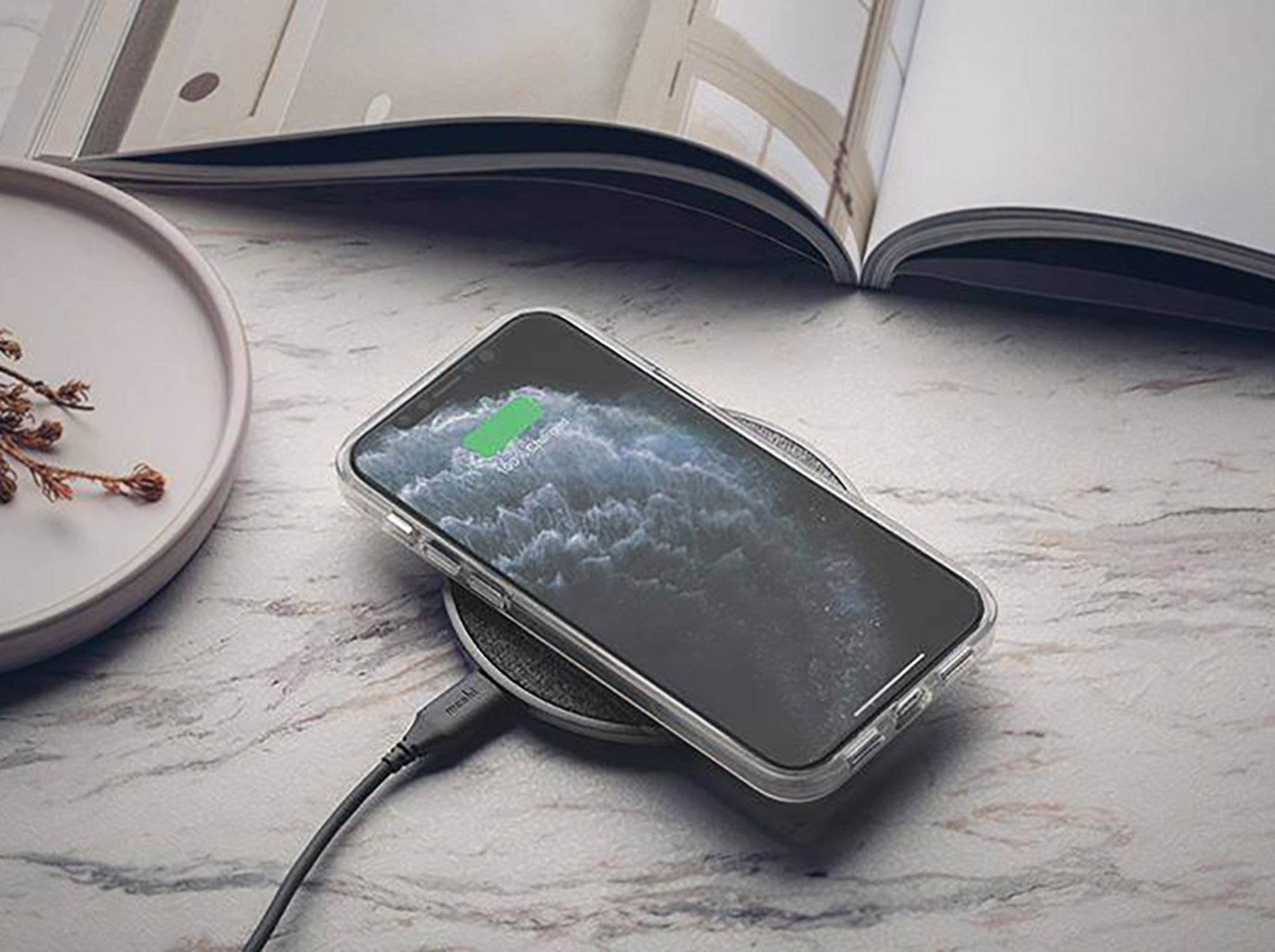 Telefon auf kabellosem Ladegerät, transparente Hülle, Aufladen. Marmoroberfläche mit Buch und Teller an der Seite.