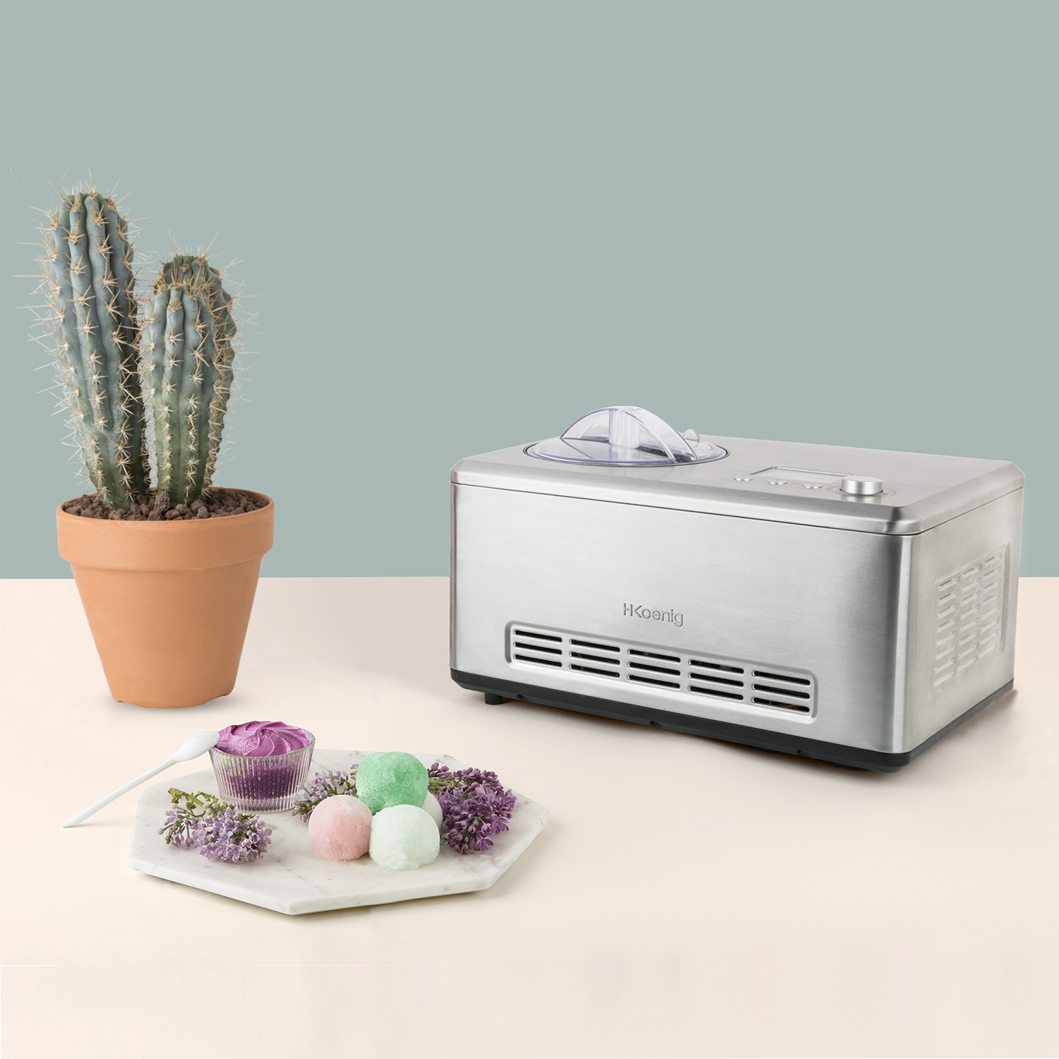 Srebrna maszyna do lodów z kaktusem, lodami i kwiatami na stole. Niebiesko-beżowe tło.