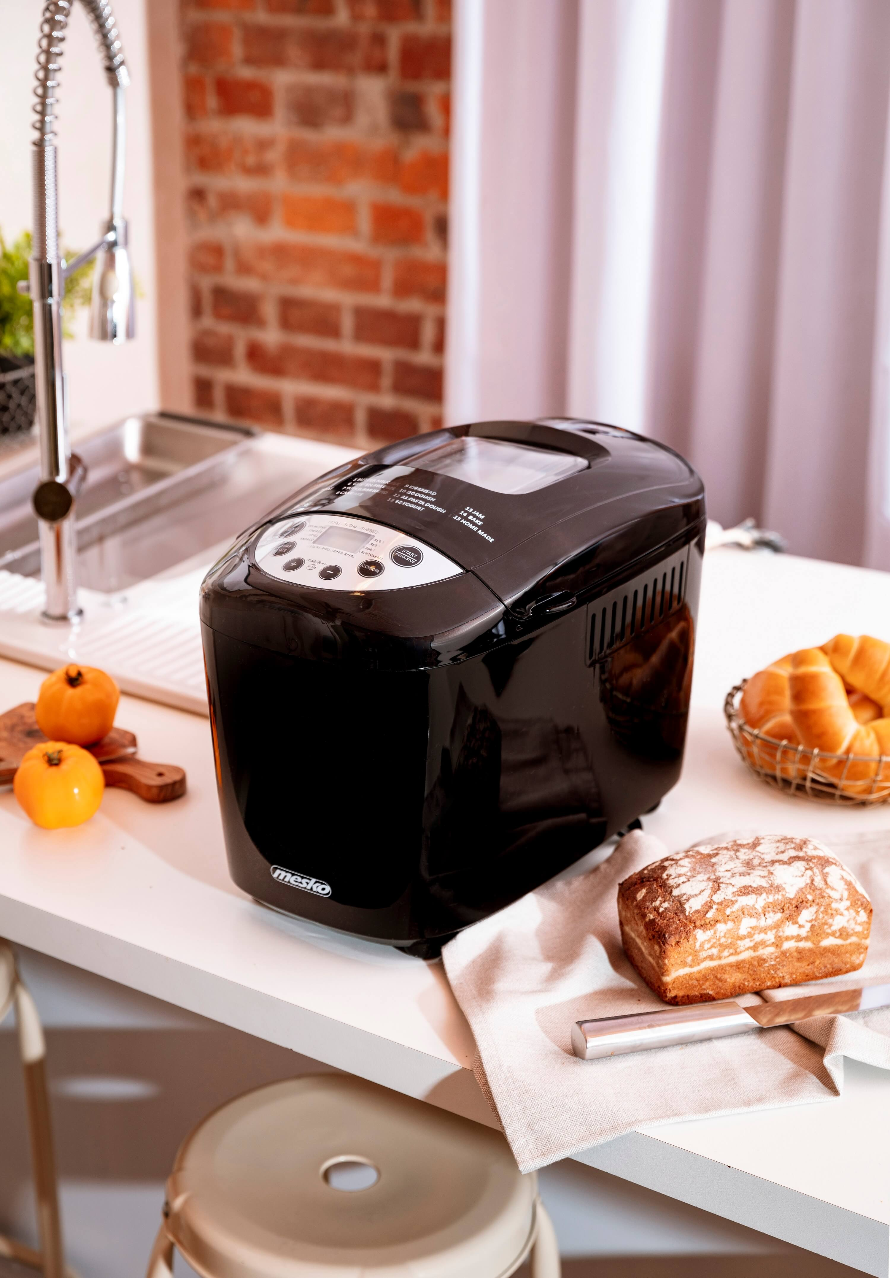 Schwarzer Brotbackautomat auf weißer Theke. Es gibt Orangen, Brot und einen Brotlaib.