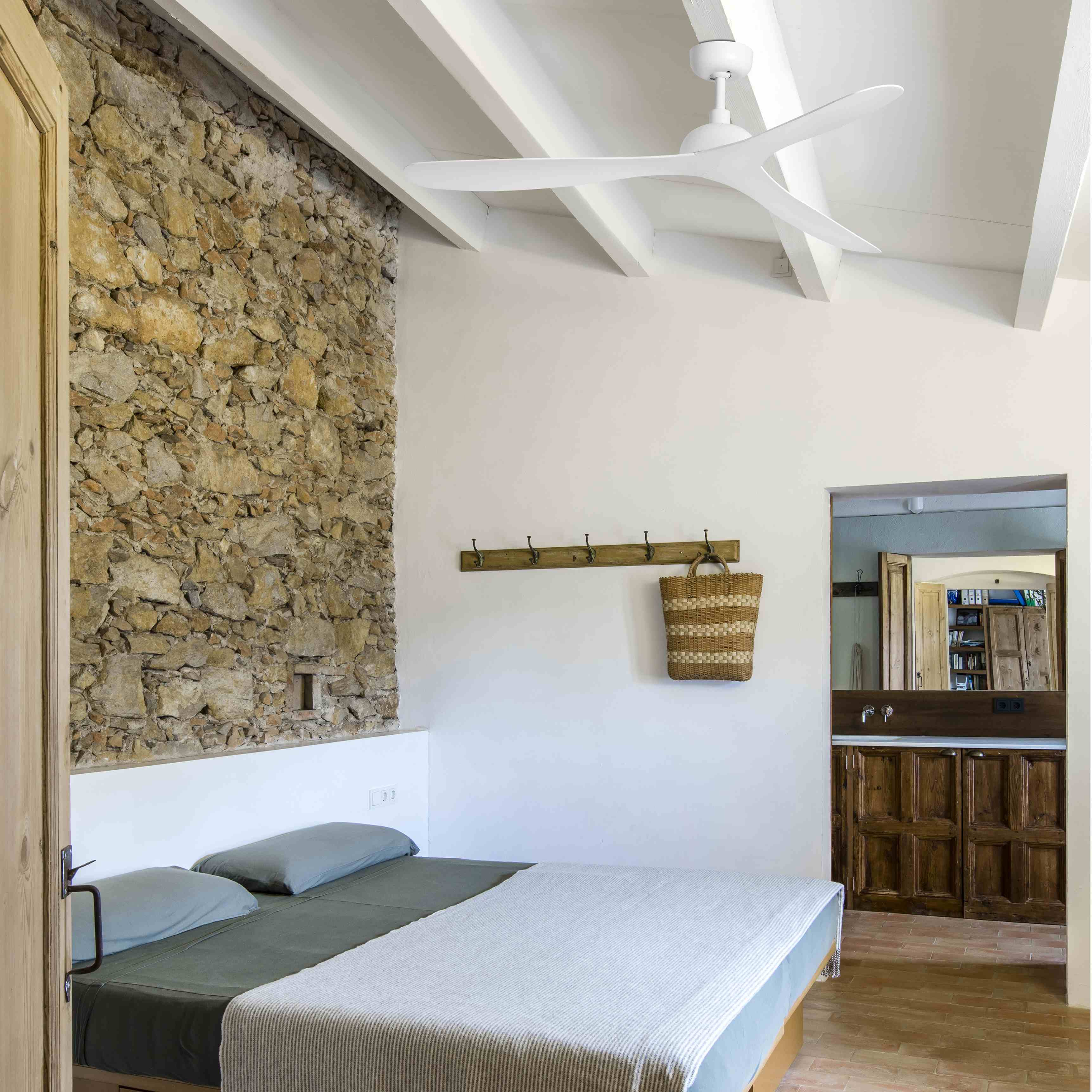 Camera da letto con muro di pietra, soffitto bianco con travi, letto con lenzuola verdi e ventilatore.