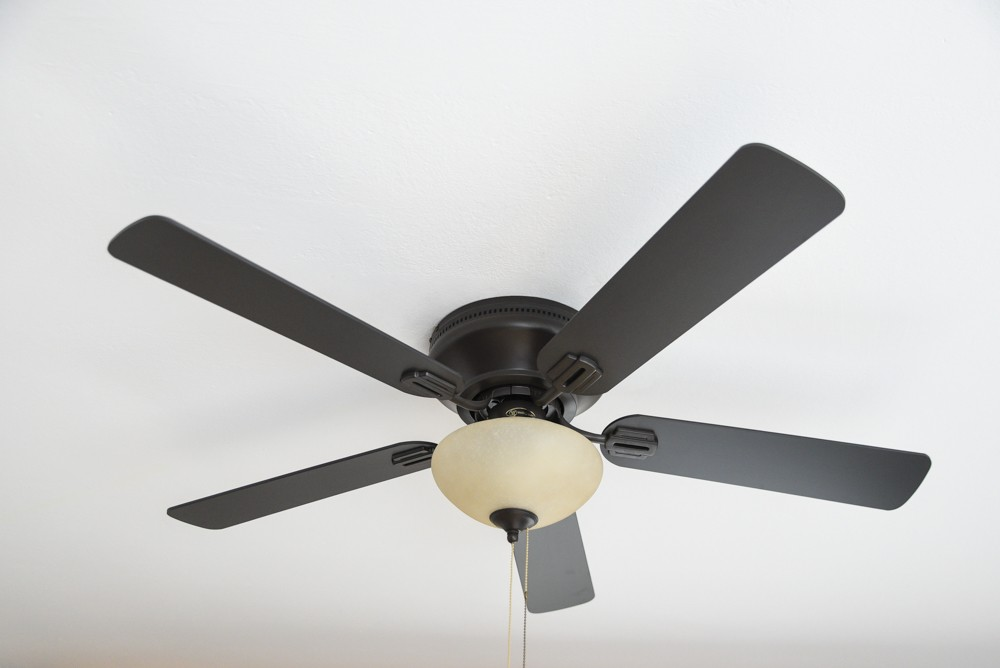 Ein Deckenventilator mit 5 schwarzen Flügeln und einer Leuchte vor weißer Decke.