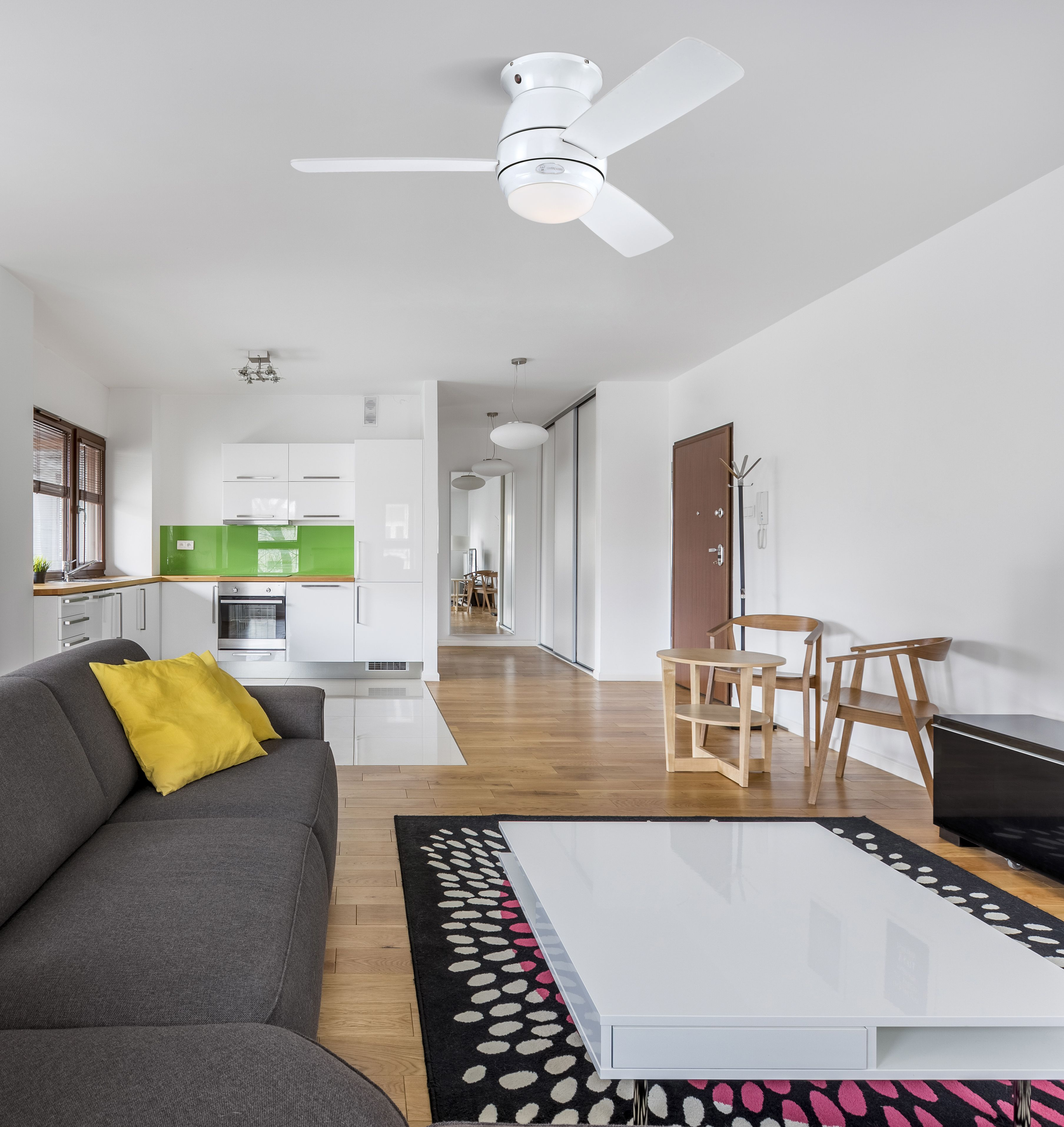 Wohnzimmer mit Deckenventilator. Graues Sofa, weißer Tisch, Holzboden, Küche im Hintergrund.