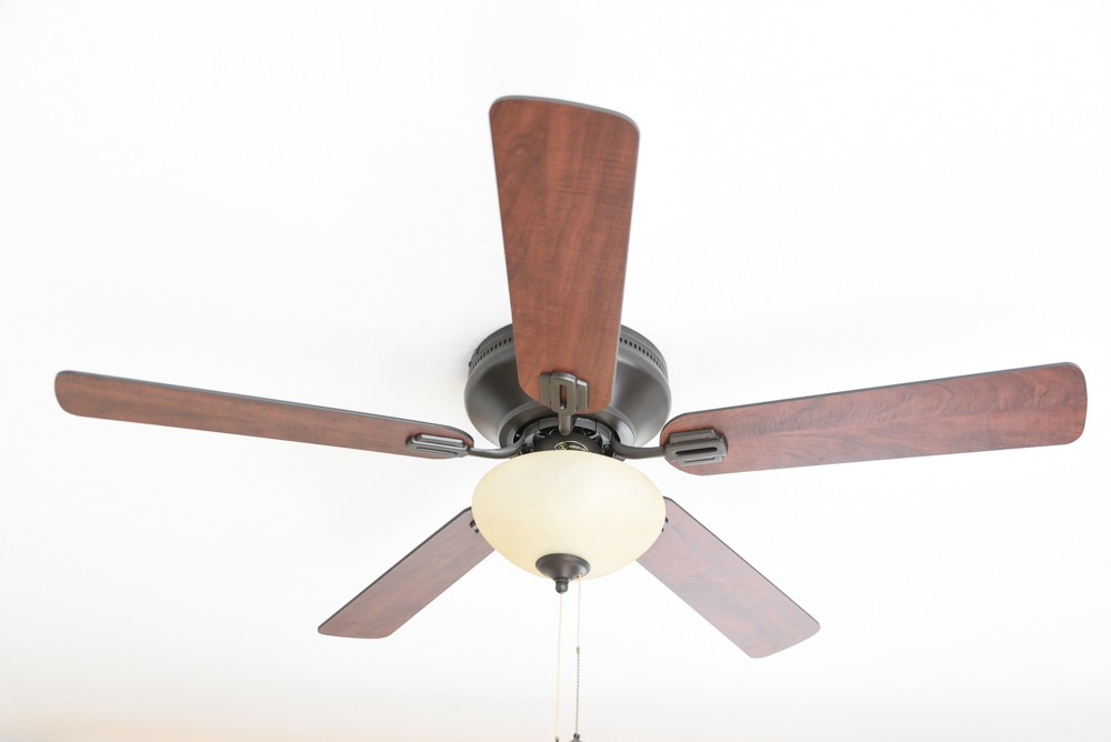 Ein Deckenventilator mit fünf Holzblättern und einer zentralen Leuchte. Weißer Hintergrund.